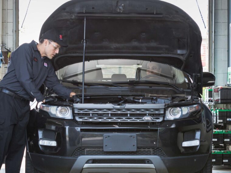 A Grease Monkey mechanic in uniform inspects under the hood of a black SUV with its hood open inside an auto repair shop, preparing for a possible clutch replacement.