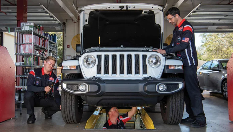 Technicians inspecting a vehicle with the hood raised, including one working underneath and one checking tire pressure.