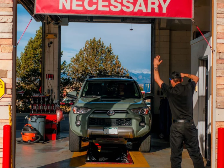 A mechanic directs a green SUV into a service bay under a red sign that reads "No Appointment Necessary," where air conditioning services and garage equipment are visible in the background.
