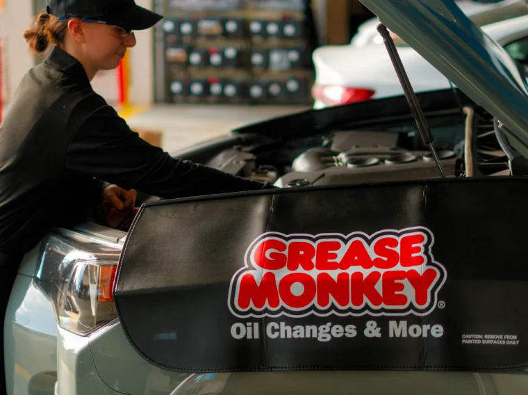 An auto technician inspects the engine of a vehicle with a "Grease Monkey Oil Changes & More" cover draped over the fender, ensuring top-notch Battery Services in a professional service garage.