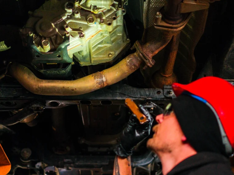 A mechanic wearing a red helmet and gloves inspects the underside of a vehicle, holding a flashlight and looking up at the car’s transmission, exhaust system, and brake services components.