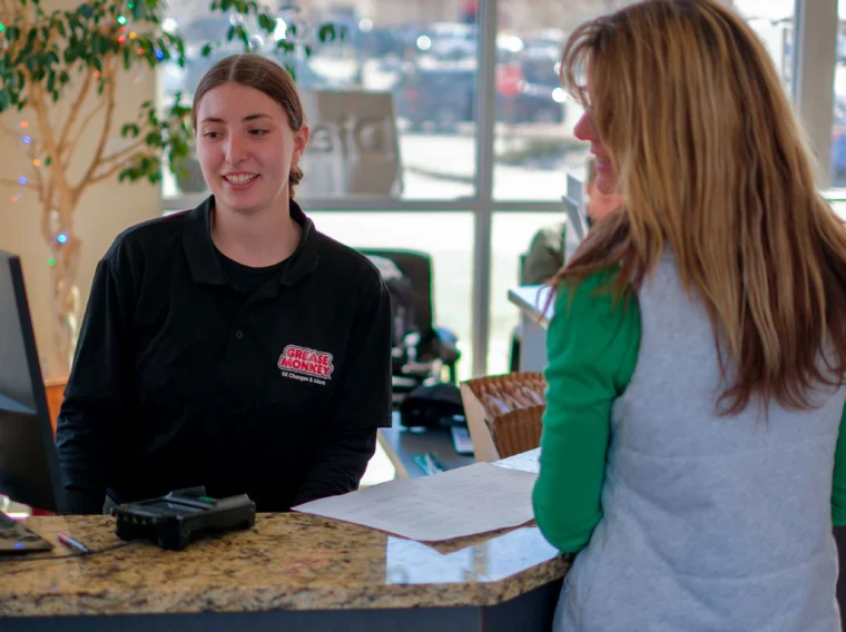 A woman stands at a reception desk talking to a receptionist about brake services, with an "OPEN" sign and large windows in the background.