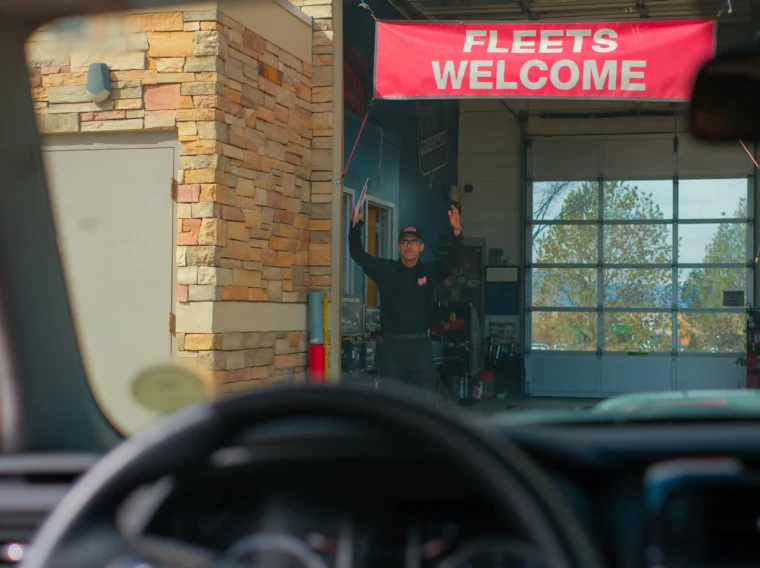A person stands under a "Fleets Welcome" sign, directing a vehicle into a stone-walled garage for car detailing, viewed from inside a car.