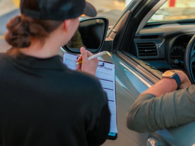A worker holding a clipboard speaks with a person in a car at a service station, while other employees handle car cleaning tasks in the background near caution tape.