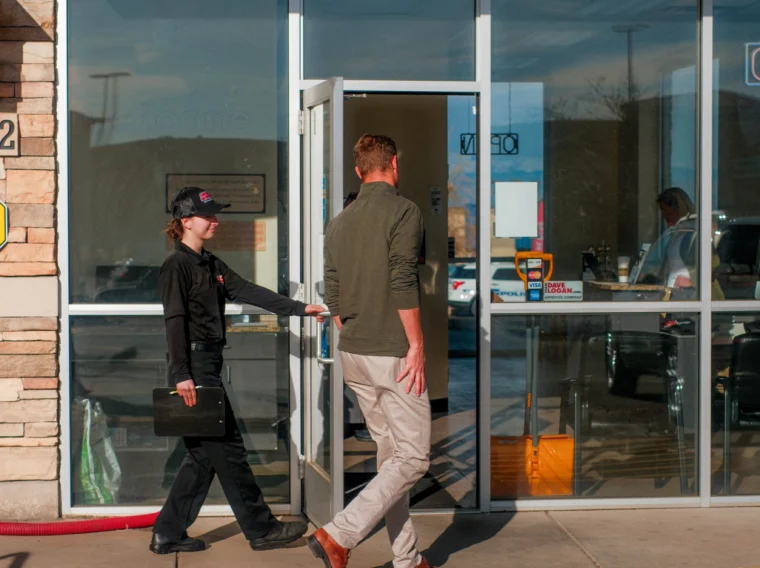 A Grease Monkey employee holds a glass door open for a man outside a building on a sunny day, discussing cylinder replacement after their visit.