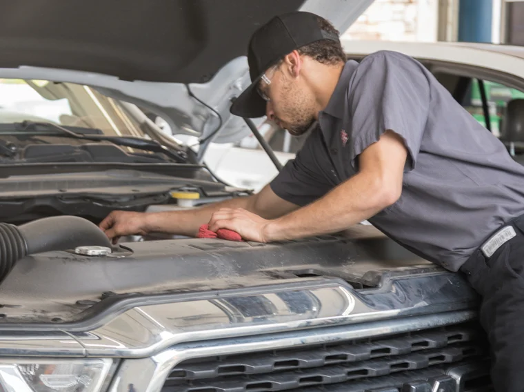 A mechanic wearing a gray uniform and cap works under the hood of a RAM truck, inspecting the engine with a red cloth in hand during a possible clutch replacement.