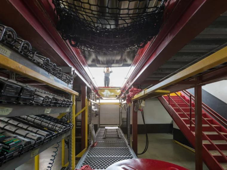 View from inside a maintenance bay showing shelves with parts for clutch replacement, a grated walkway, red stairs on the right, and a person standing above an open hatch at the far end.
