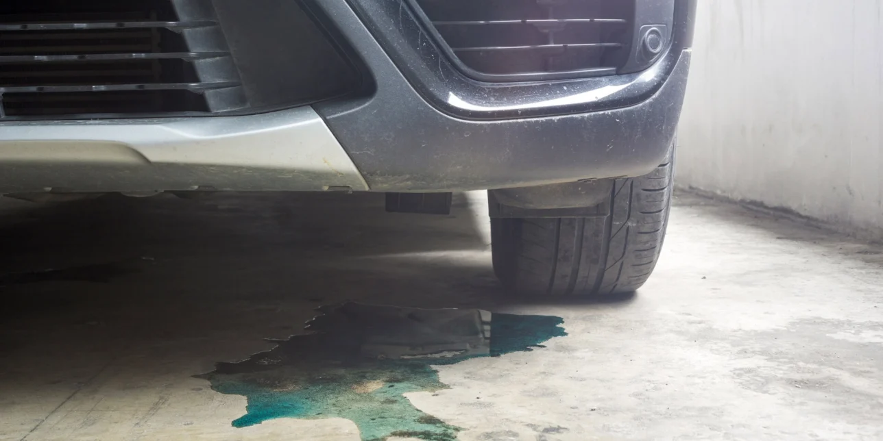 A close-up of a car's front bumper and tire shows a greenish fluid leaking onto the concrete floor—one of the common car leaks that highlights the need for regular car maintenance.