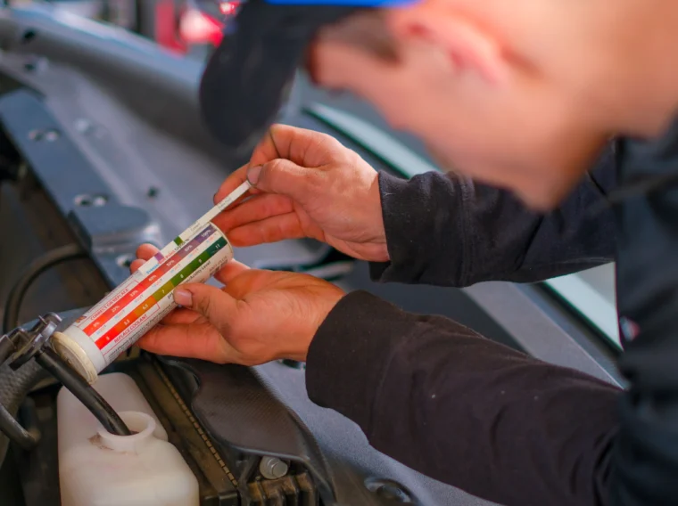 A person in a cap comparing the color of engine coolant near a white reservoir during a cooling system repair.