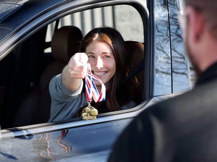 A woman sitting in a car smiles and holds out a gold medal with a ribbon toward a person standing outside the vehicle.