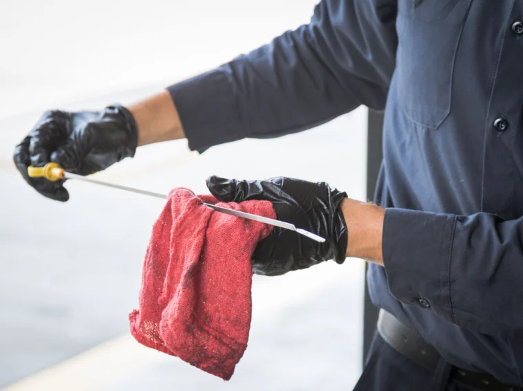 A person wearing black gloves wipes an engine oil dipstick with a red cloth, likely performing vehicle maintenance as part of routine engine services.