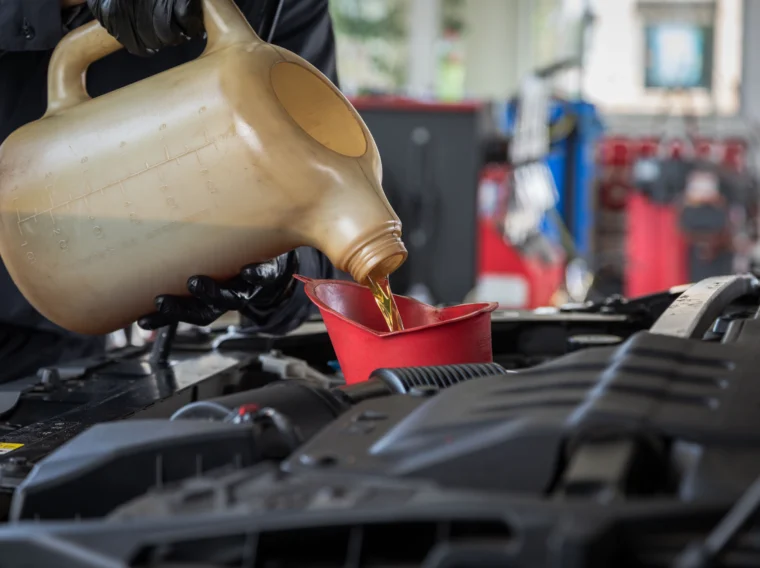 A person pours motor oil from a large container into a red funnel positioned in a car engine bay while performing engine services at an automotive service shop.