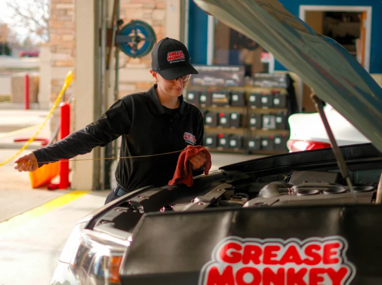 A mechanic in a Grease Monkey uniform checks the oil level under the hood of a vehicle, showcasing expert Engine Services inside an auto service garage.