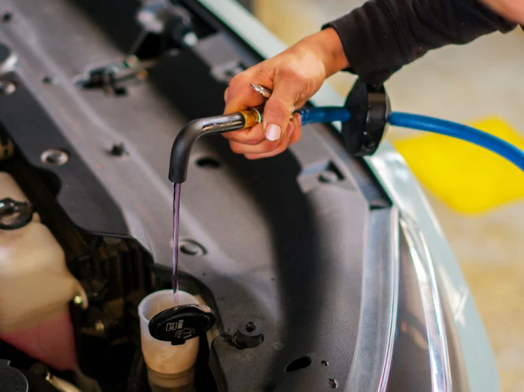 A person pours coolant into a car's windshield washer fluid’s reservoir using a hose, with the hood open in a garage setting—a common step in routine Fluid Exchange Services.