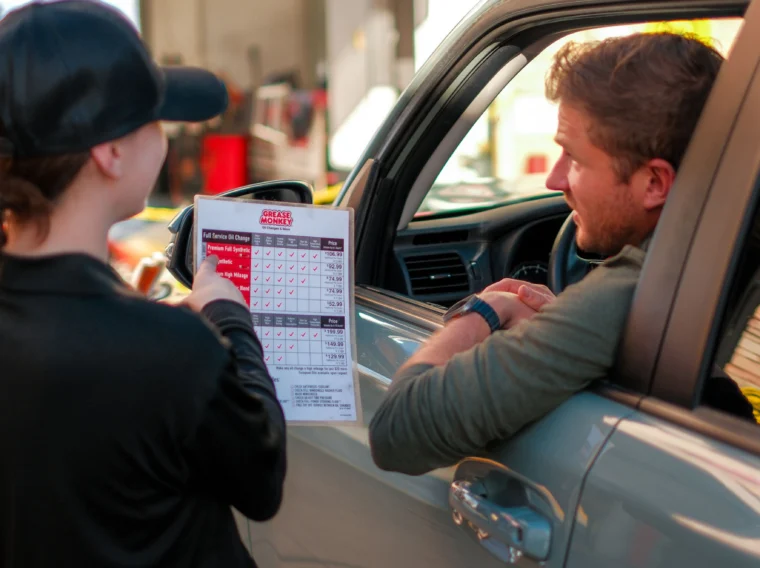 A person in uniform shows a checklist to a man in his car at a drive-through service location specializing in Fluid Exchange Services.