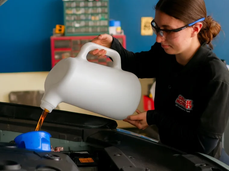 A person wearing safety glasses pours oil from a large container into a car engine using a blue funnel.