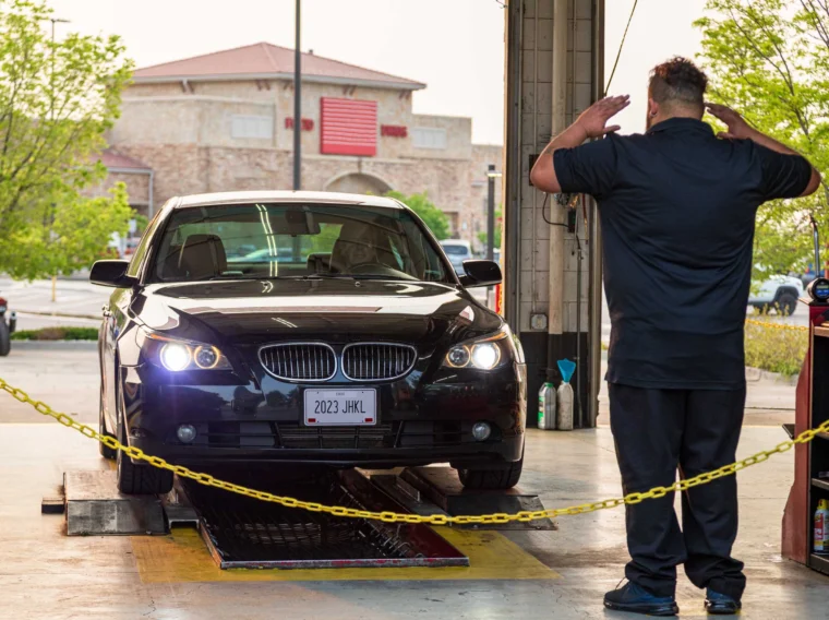 A person in black signals a black BMW with headlights on to move forward inside an auto service garage.