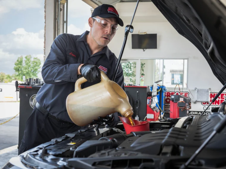 A mechanic wearing safety glasses and gloves pours engine oil into a car’s engine using a large container at an auto service center.