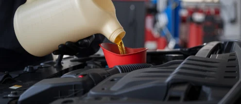 A person wearing black gloves pours engine oil from a large container into a red funnel placed in a car engine bay.