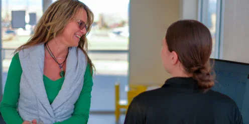 A woman wearing glasses and a green shirt smiles while speaking to another woman with brown hair in a bun, indoors near a window.